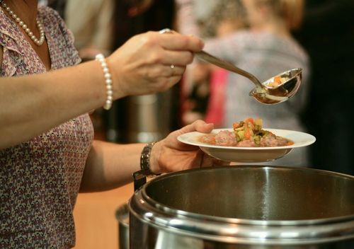 Ein Teller wird über einen großen Topf gehalten, aus den mit einer Kelle Essen geschöpft wird.